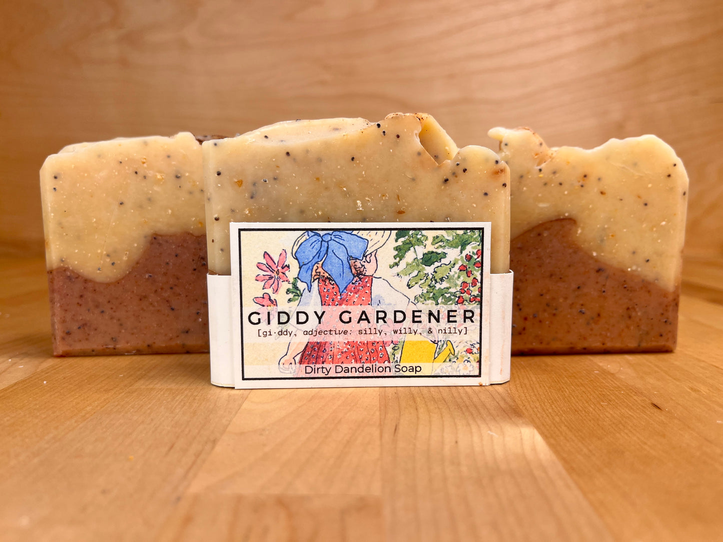 Front-facing view of a handmade Giddy Gardener soap bar wrapped with an illustrated label, flanked by two unwrapped bars on a wooden surface, showing a reddish-brown base and a speckled golden-yellow top layer with a softly textured edge