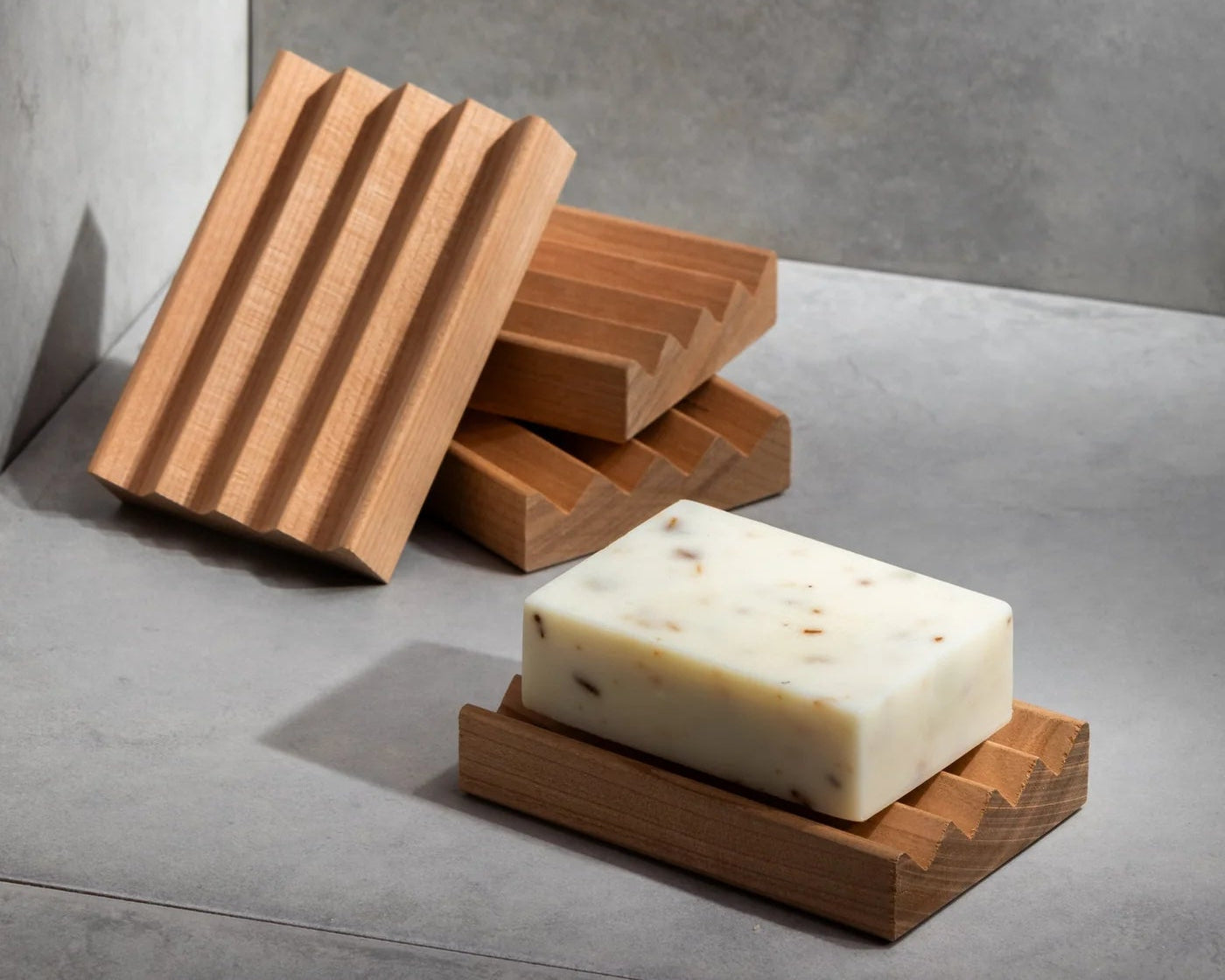 A wooden soap dish with a minimalist design, featuring wide angled channels, placed on a grey surface with a block of soap resting on top.