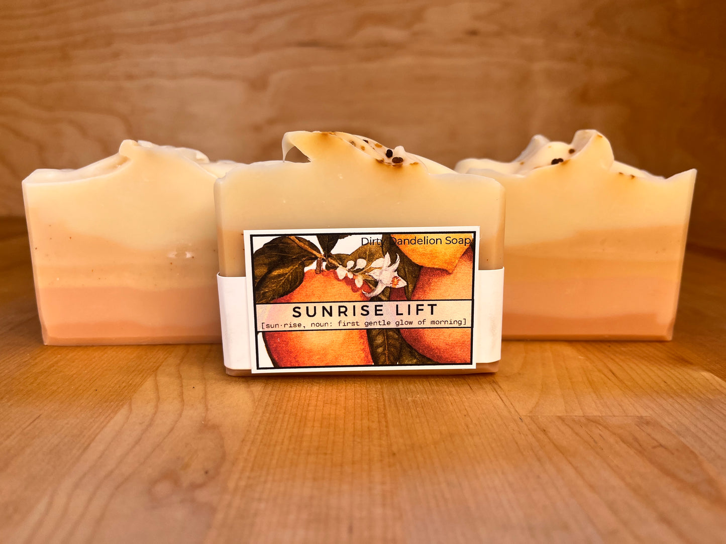 Sunrise Lift soap bars displayed upright on a wooden surface, showing soft horizontal layers of peach, golden yellow, and cream, with a centered wrapped bar featuring the Sunrise Lift illustrated label and gently textured tops with small botanical accents