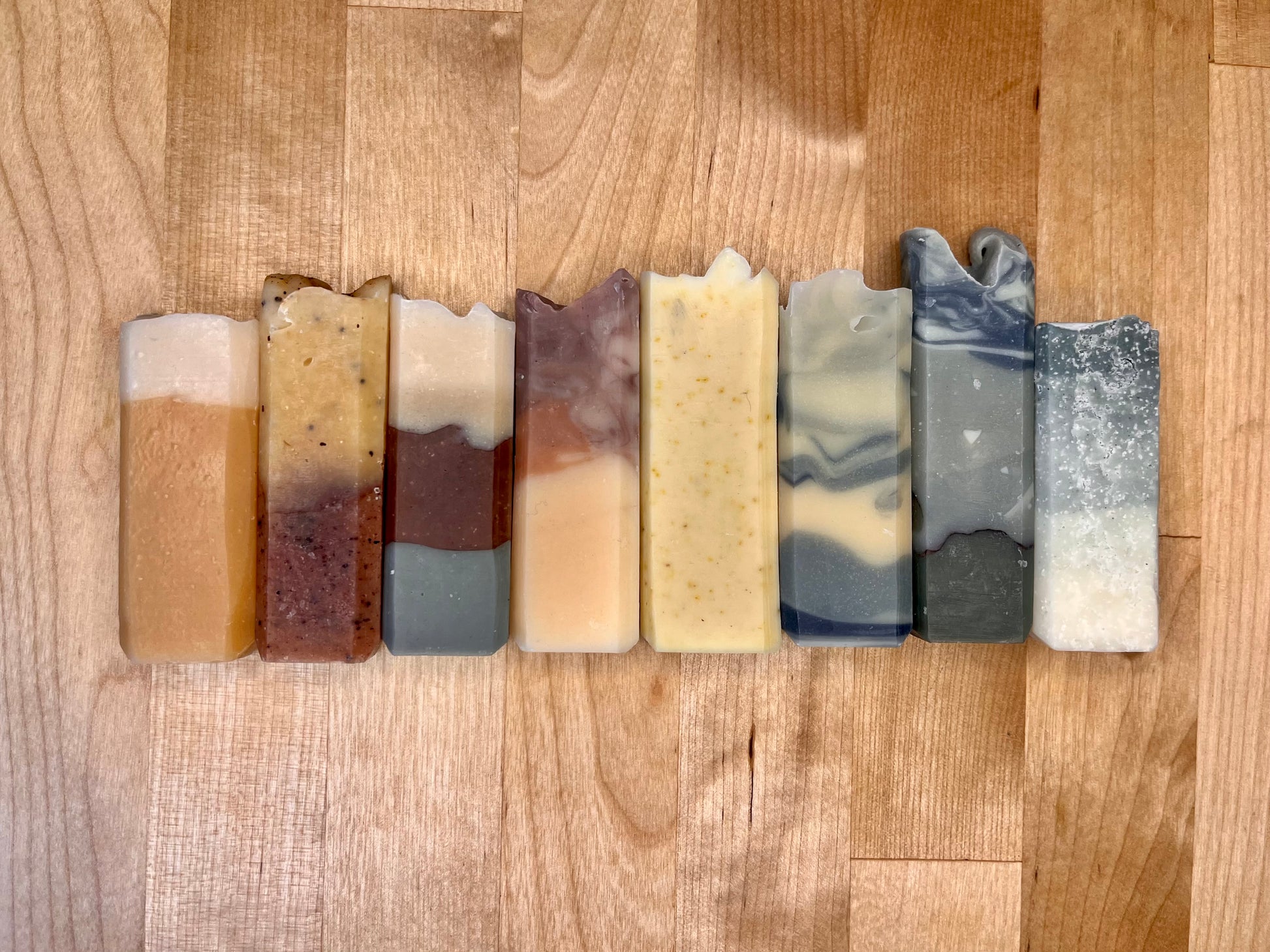 Row of eight narrow handmade soap bars standing upright on a wooden surface, each with a distinct color pattern including layered amber and cream, speckled yellow, marbled gray, and blue-gray swirl designs.