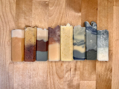 Row of eight narrow handmade soap bars standing upright on a wooden surface, each with a distinct color pattern including layered amber and cream, speckled yellow, marbled gray, and blue-gray swirl designs.