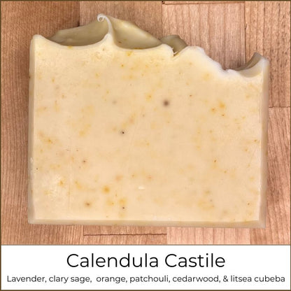 Single handmade cold process soap bar with a pale cream color and small botanical speckles throughout, finished with a softly sculpted top edge, photographed on a wooden surface and labeled “Calendula Castile.”