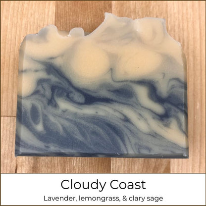 Single handmade cold process soap bar labeled “Cloudy Coast,” featuring soft cream and slate-gray marbling with flowing, wave-like swirls and a gently sculpted top edge, photographed on a wooden surface.