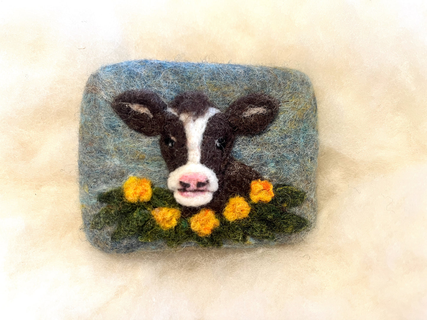 Hand-felted Wooly Washer soap featuring a brown and white goat face with pink nose, surrounded by yellow flowers and green grass on a soft blue background, photographed on a light wool surface.
