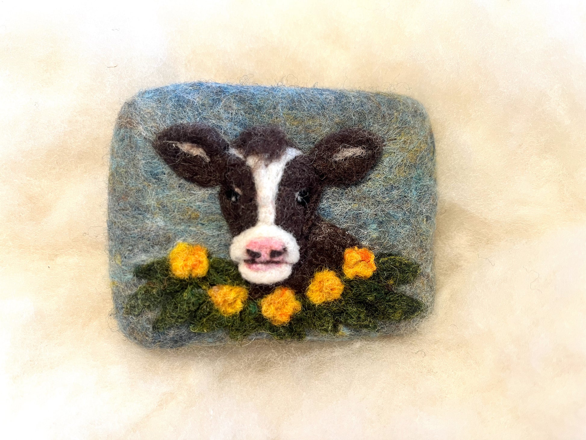 Hand-felted Wooly Washer soap featuring a brown and white goat face with pink nose, surrounded by yellow flowers and green grass on a soft blue background, photographed on a light wool surface.