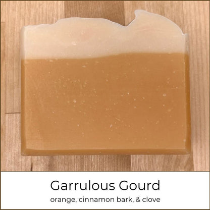 Single handmade cold process soap bar with a warm amber body and a smooth creamy white top, photographed on a wooden surface and labeled “Garrulous Gourd.”