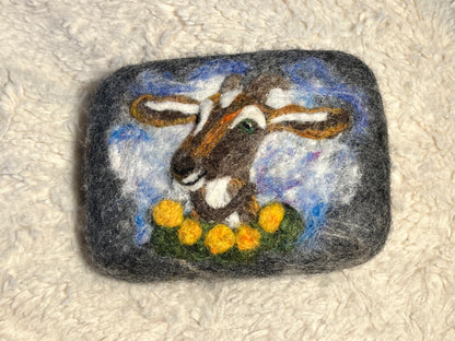 Needle-felted Goat Wooly Washer soap with a dark gray wool cover and a hand-felted goat face in brown, white, blue, and yellow fiber, photographed on a cream textured surface.