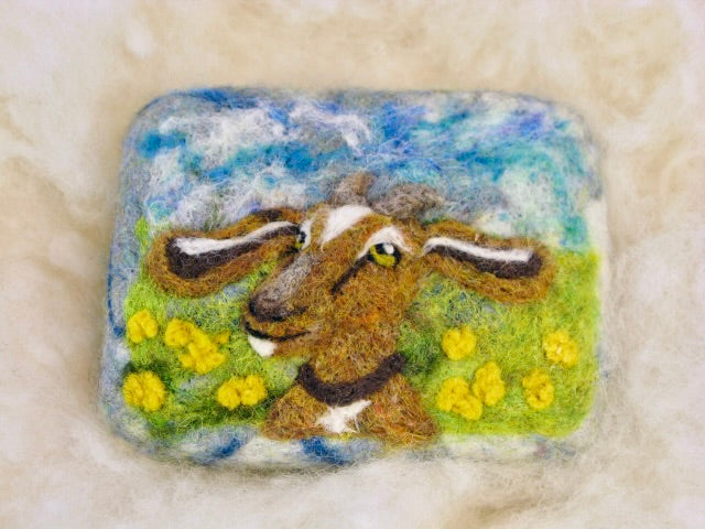 Needle-felted wooly washer soap with a soft gray wool cover featuring a hand-felted goat portrait in brown and white fiber, set against a green meadow with yellow flowers and a blue sky background, photographed on a cream wool surface.