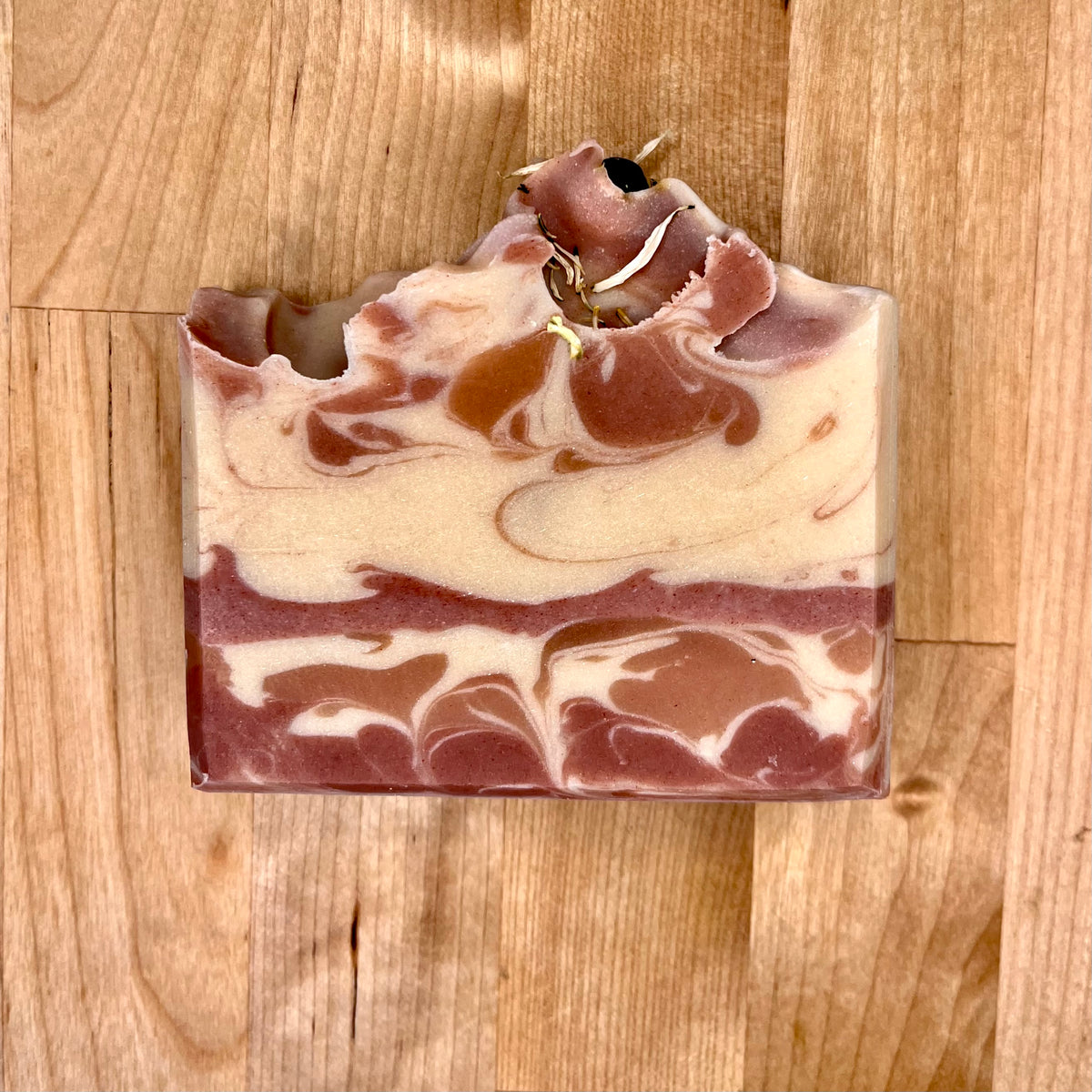 Single Happy Holly cold process soap bar on a wooden surface, featuring marbled cream and rose-toned swirls, a textured piped top, and small botanical accents embedded along the top edge