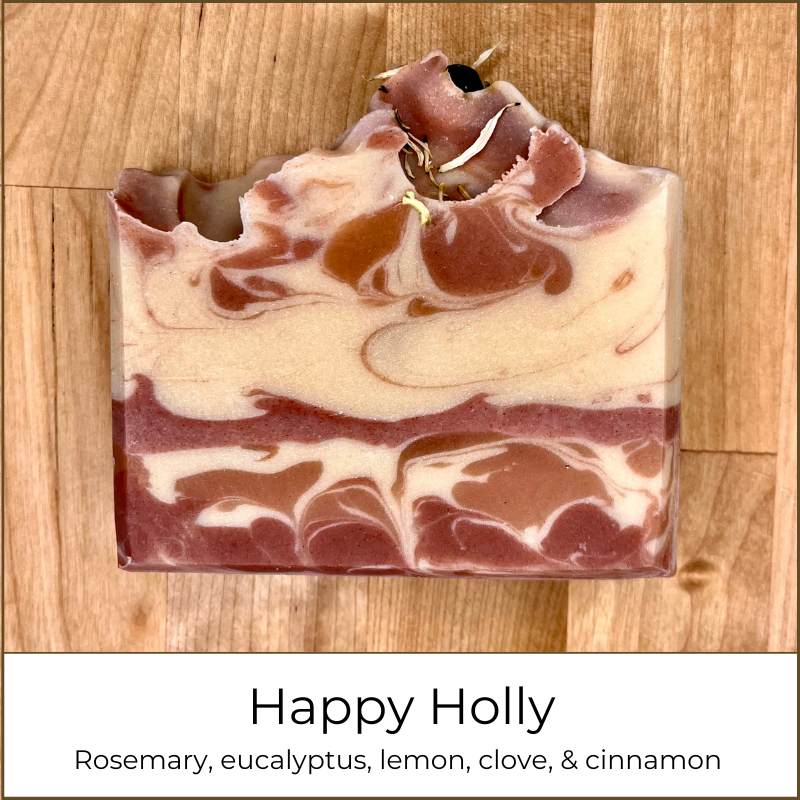Happy Holly cold process soap bar photographed on a wooden surface, showing layered cream and rose-colored swirls with a textured top, topped with small botanical pieces, and labeled with essential oils rosemary, eucalyptus, lemon, clove, and cinnamon