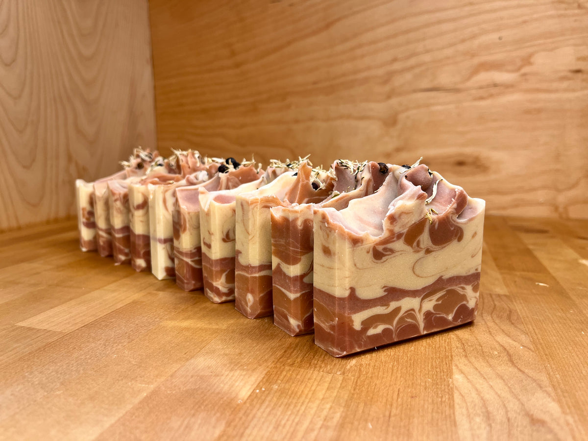 Happy Holly cold process soap bars lined up on a wooden surface, showing marbled cream and rose-toned swirls, textured piped tops, and subtle botanical details along the edges