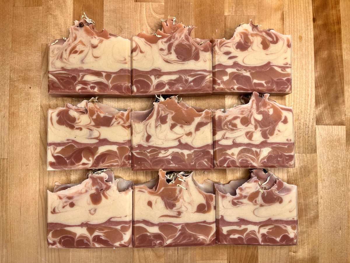 Nine Happy Holly cold process soap bars arranged in a grid on a wooden surface, showing creamy ivory and deep red swirls with textured tops and botanical accents