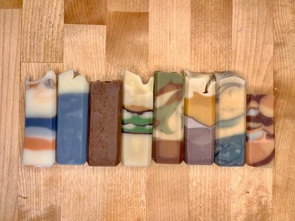 Row of handmade cold-process soap bars arranged side by side on a wooden surface, each with layered colors and marbled patterns in creams, blues, greens, browns, and warm earth tones.