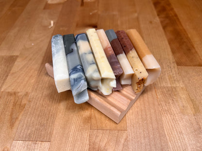 Stack of eight narrow handmade soap bars with varied colors and patterns, including marbled gray, speckled yellow, layered cream and brown, and amber tones, resting on a small wooden soap dish on a wooden surface.