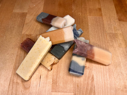 Scattered assortment of narrow handmade soap bars with mixed colors and layered patterns, including amber, cream, marbled gray, speckled yellow, and reddish-brown tones, spread across a wooden surface.