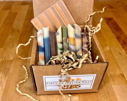 Open kraft box containing assorted handmade soap bars arranged upright with crinkle paper, labeled Dirty Dandelion Soap.