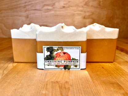 Three handmade cold process soap bars with warm amber bases and creamy white tops, one wrapped with a pumpkin-themed “Smashing Pumpkin” Dirty Dandelion Soap label, displayed on a wooden surface.