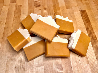 Several rectangular handmade soap bars stacked and scattered on a wooden table, each with a light top layer and a darker tan base.
