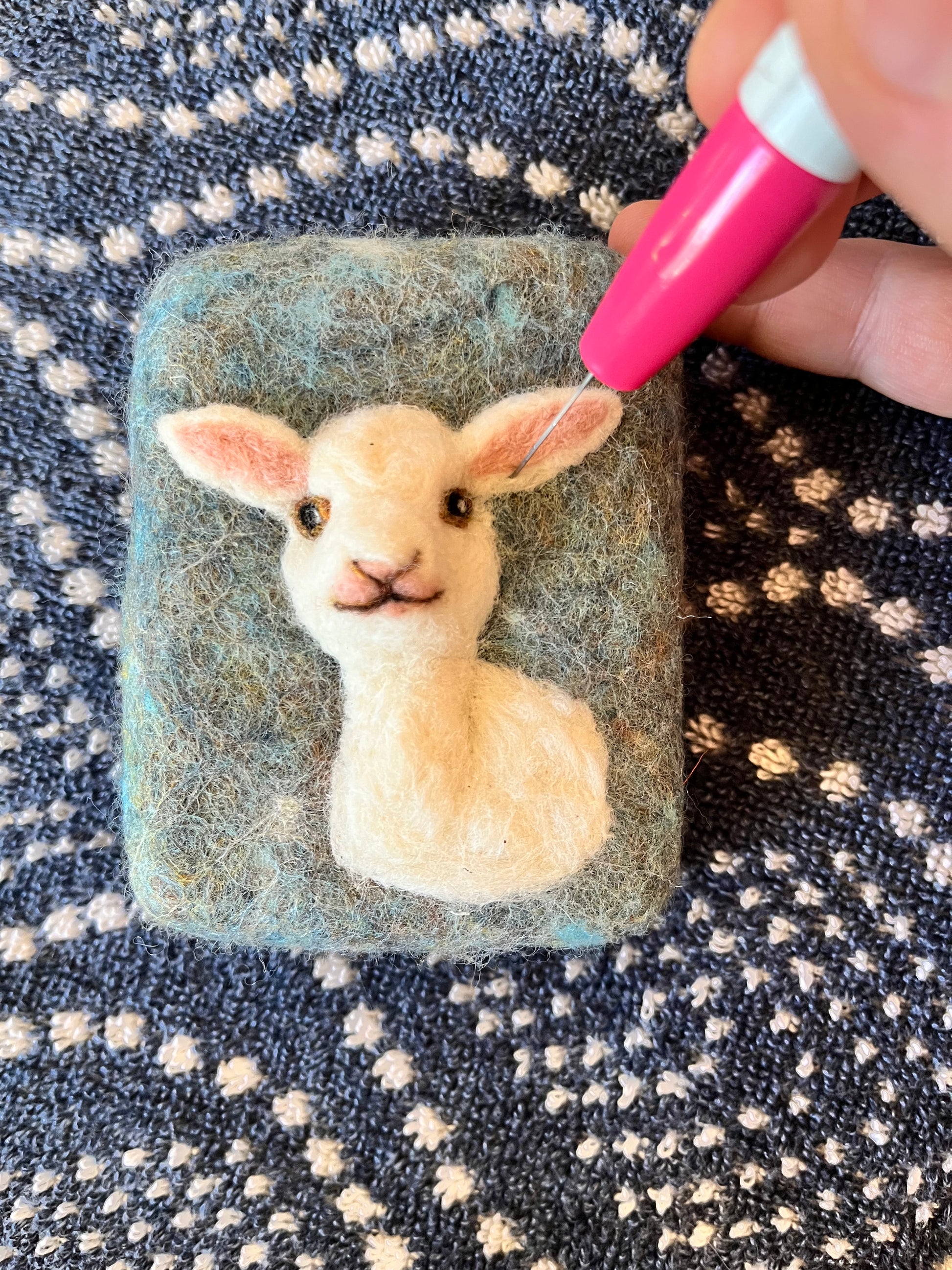 Hand-felted Wooly Washer soap featuring a white lamb with pink inner ears and stitched facial details on a soft blue-gray background, shown mid-process with a felting needle shaping the ear.
