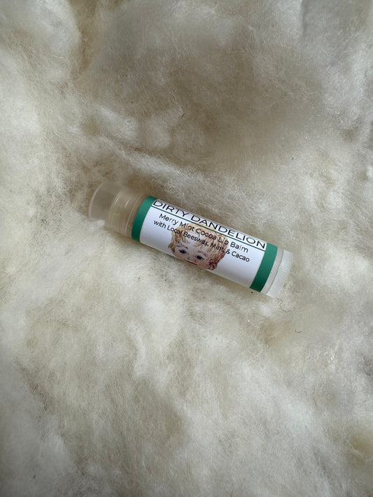 Single lip balm tube labeled “Merry Mint Cocoa Balm” from Dirty Dandelion, with a white label, green accent bands, and illustrated face graphic, resting on a soft, cream-colored wool background.