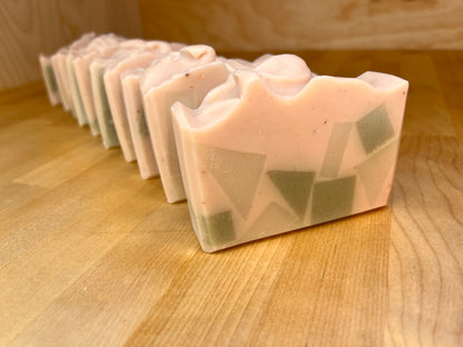 A row of Mosaic Meadow soap bars with a pale pink base and embedded green geometric shapes, lined up on a wooden surface and photographed at an angle