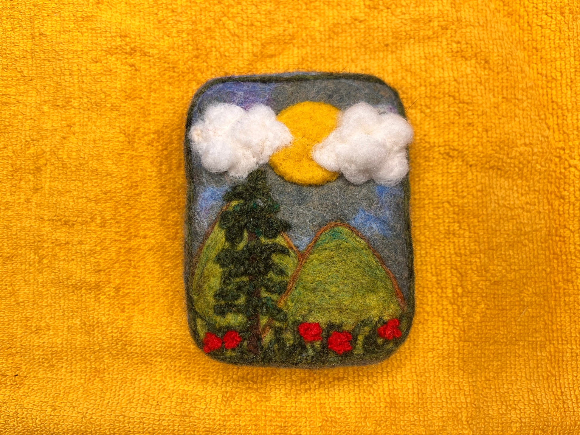 Handmade Wooly Washer felted soap featuring a needle-felted landscape scene with green hills, a pine tree, red flowers, white clouds, and a yellow sun against a gray-blue sky, photographed on a yellow towel.