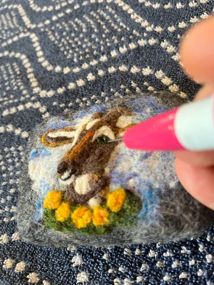 Close-up of a needle-felted Wooly Washer soap in progress, showing a hand using a felting needle to add detail to a goat’s eye on a wool design with yellow flowers and a blue sky background.
