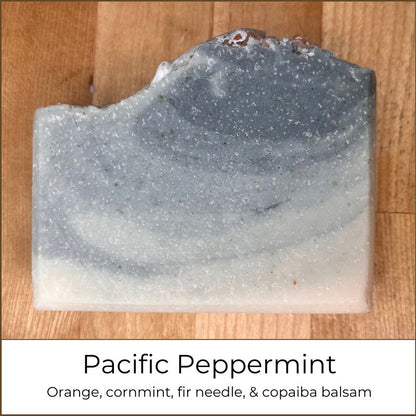Single handmade cold process soap bar labeled “Pacific Peppermint,” featuring layered pale cream and cool blue-gray tones with fine speckling and a softly curved, uneven top edge, photographed on a wooden surface.