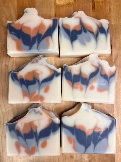 Six handmade soap bars arranged in a grid, featuring blue-gray and clay-colored swirls on a creamy base.