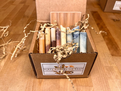 Open cardboard gift box containing a curated sampler of narrow handmade soap bars in varied colors, arranged upright with shredded paper packing and a Dirty Dandelion Soap label on the front, placed on a wooden surface.