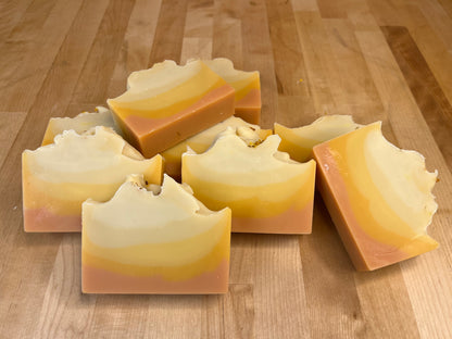 Sunrise Lift soap bars stacked and angled on a wooden surface, showing smooth curved layers of cream, pale yellow, golden orange, and blush pink with softly sculpted tops