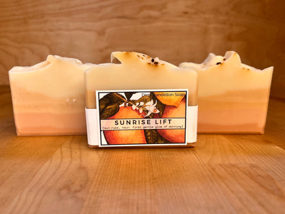 Sunrise Lift soap bars displayed upright on a wooden surface, showing soft horizontal layers of peach, golden yellow, and cream, with a centered wrapped bar featuring the Sunrise Lift illustrated label and gently textured tops with small botanical accents
