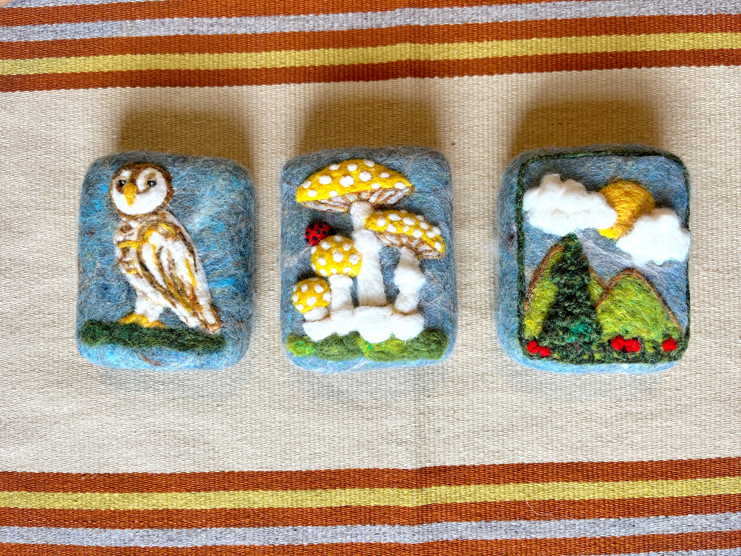 Three handmade Wooly Washer felted soaps displayed side by side, featuring needle-felted designs of an owl perched on a branch, yellow spotted mushrooms with a ladybug, and a landscape with green hills, clouds, and a yellow sun.
